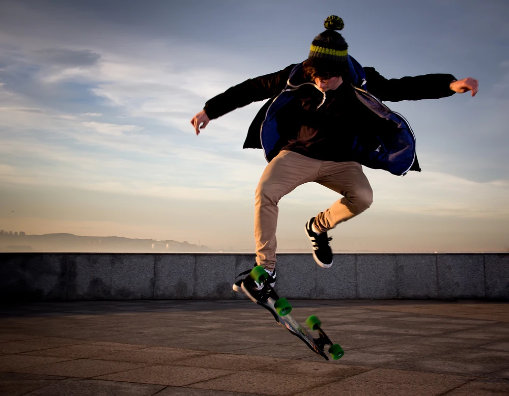 A guy in a jacket and light jeans jumps on a skateboard