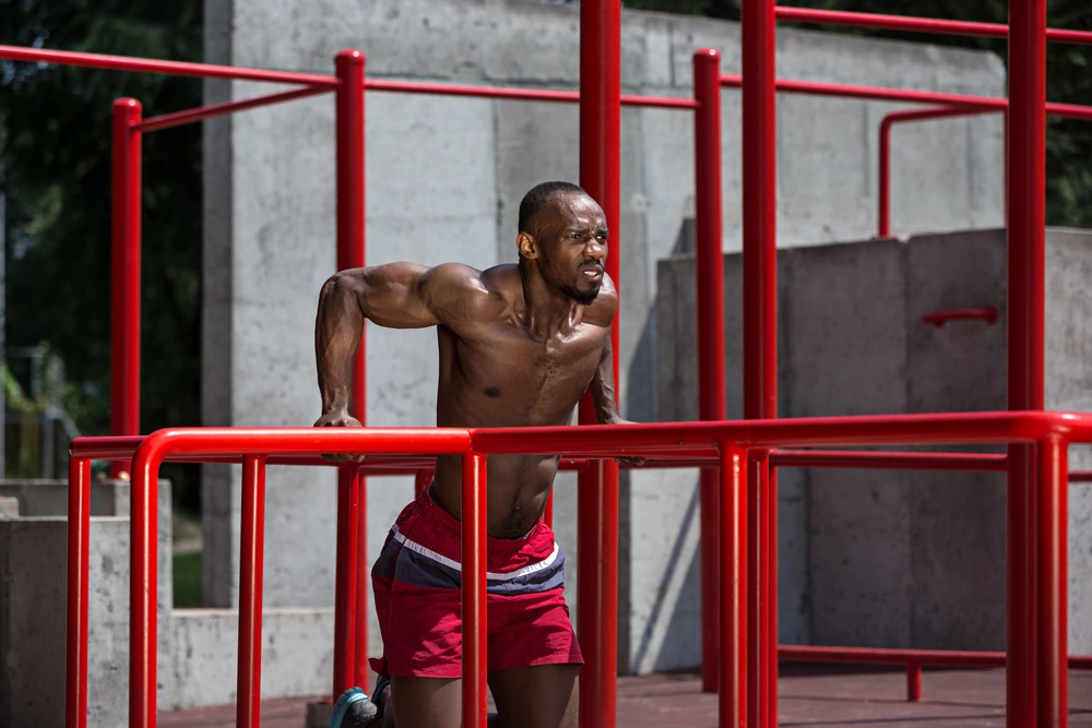 A guy in red shorts without a T-shirt does push-ups on the parallel bars